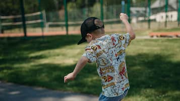 Un bambino gioca a campana sulla scuola in un parco giochi con una recinzione verde e alberi sullo sfondo.