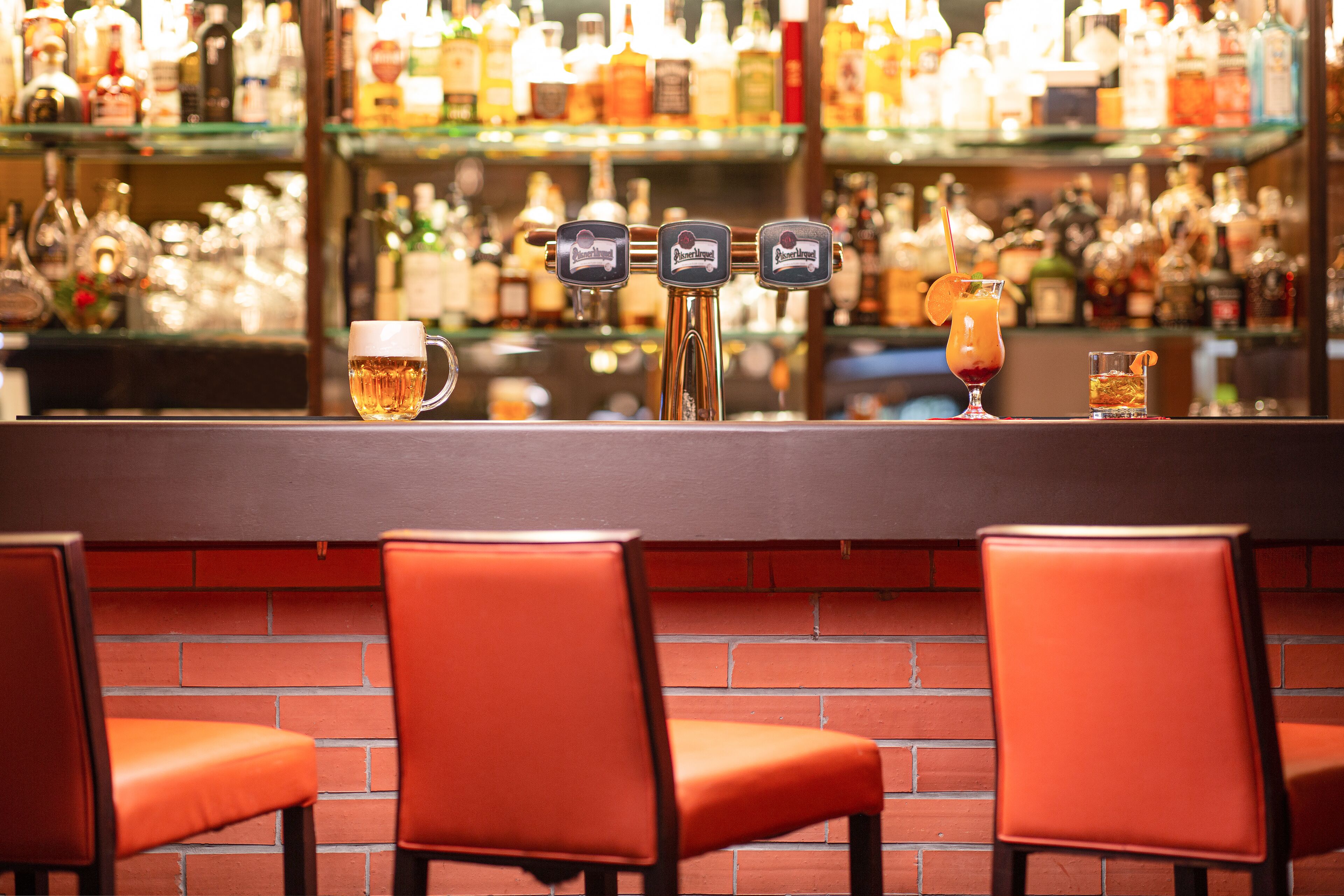 Un bar avec un mur en briques, trois tabourets et une variété de bouteilles et de verres disposés sur des étagères.
