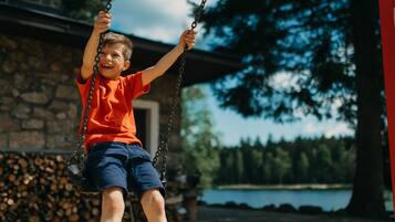 Een kind schommelt voor een stenen gebouw, met een meer en bomen op de achtergrond.