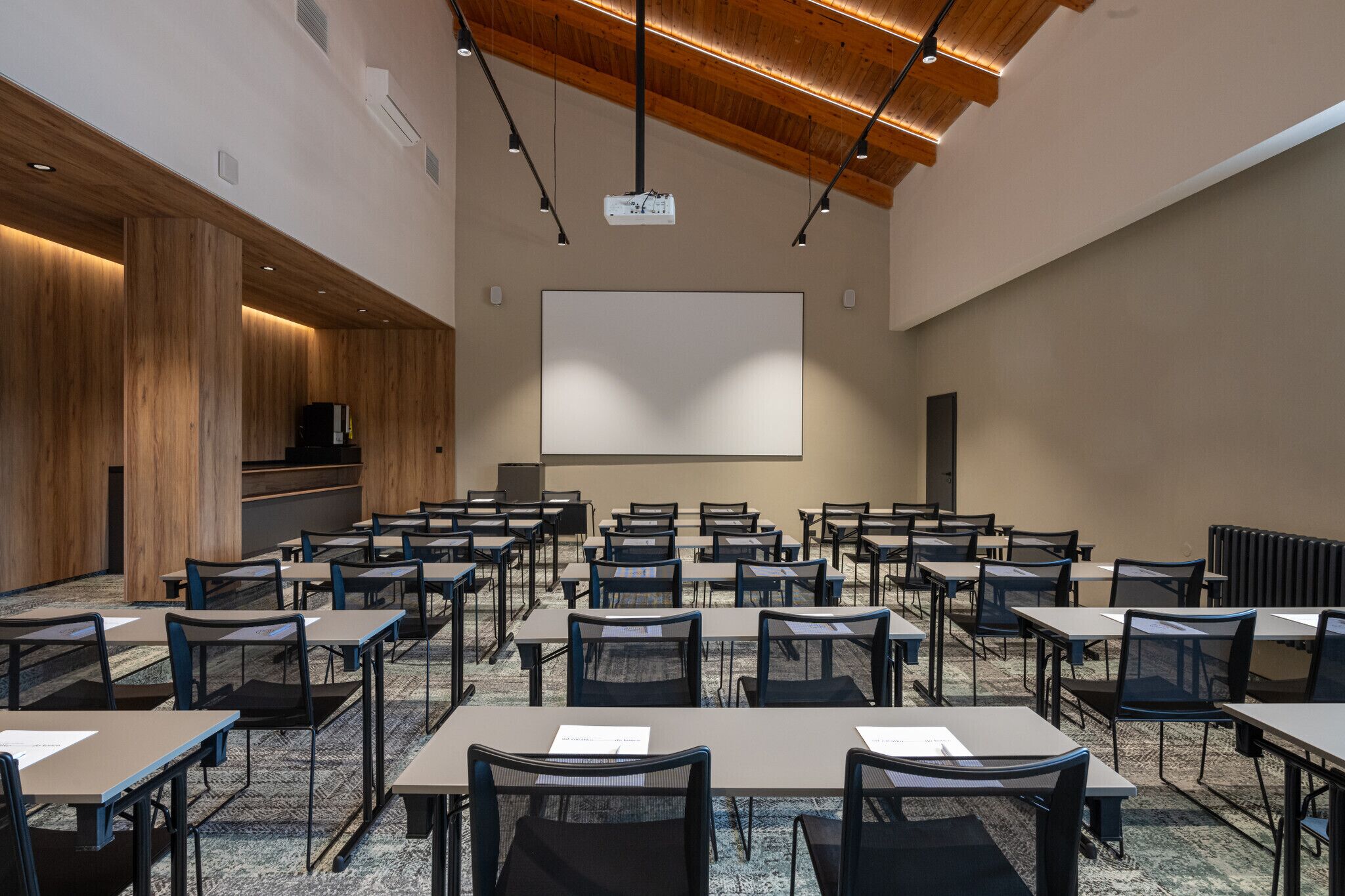 Un salón de conferencias con varias mesas y sillas, una pantalla grande y techo de madera.