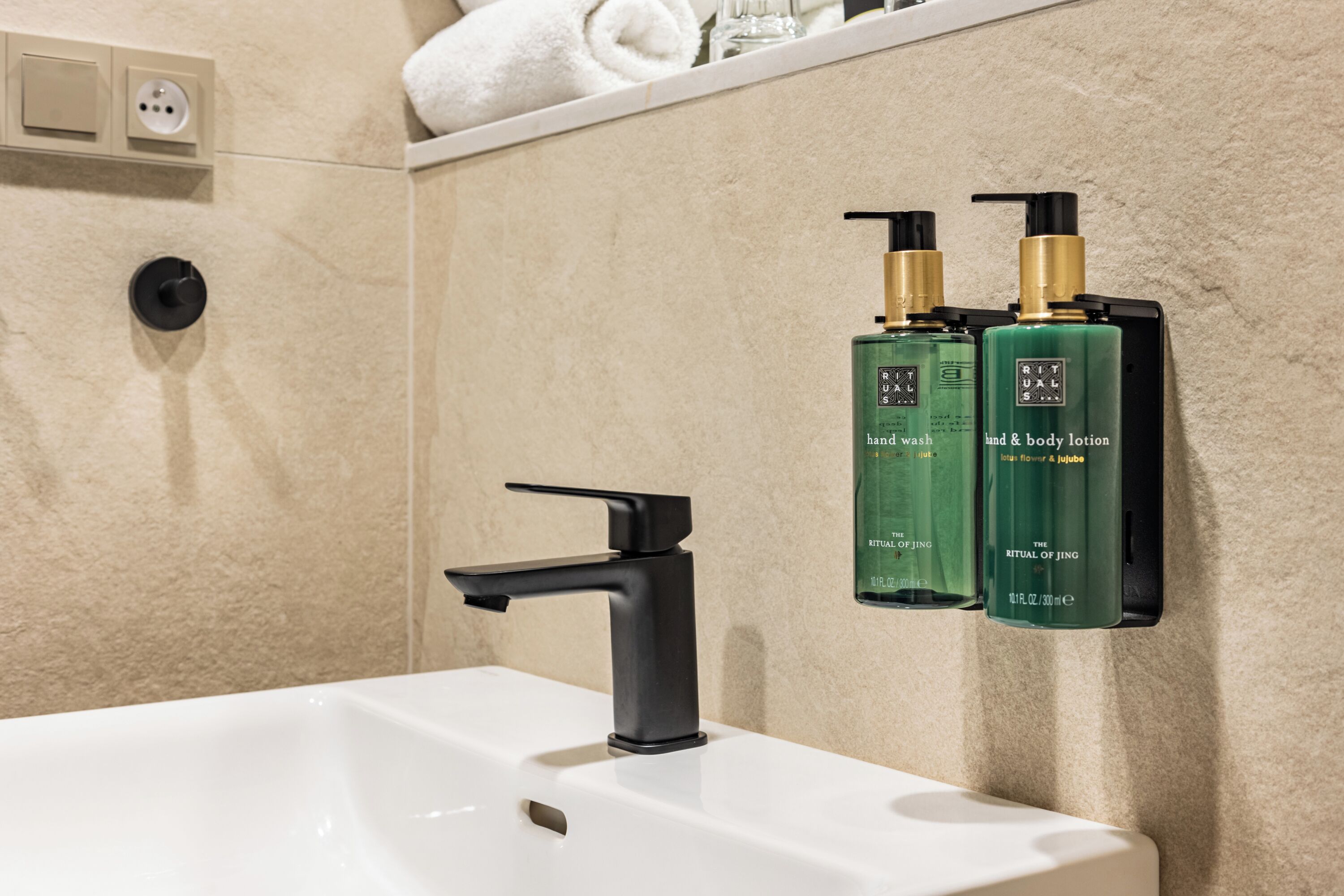 Un baño con lavamanos blanco, grifo negro y dos botellas verdes de jabón de manos y loción corporal Rituals of Jing.