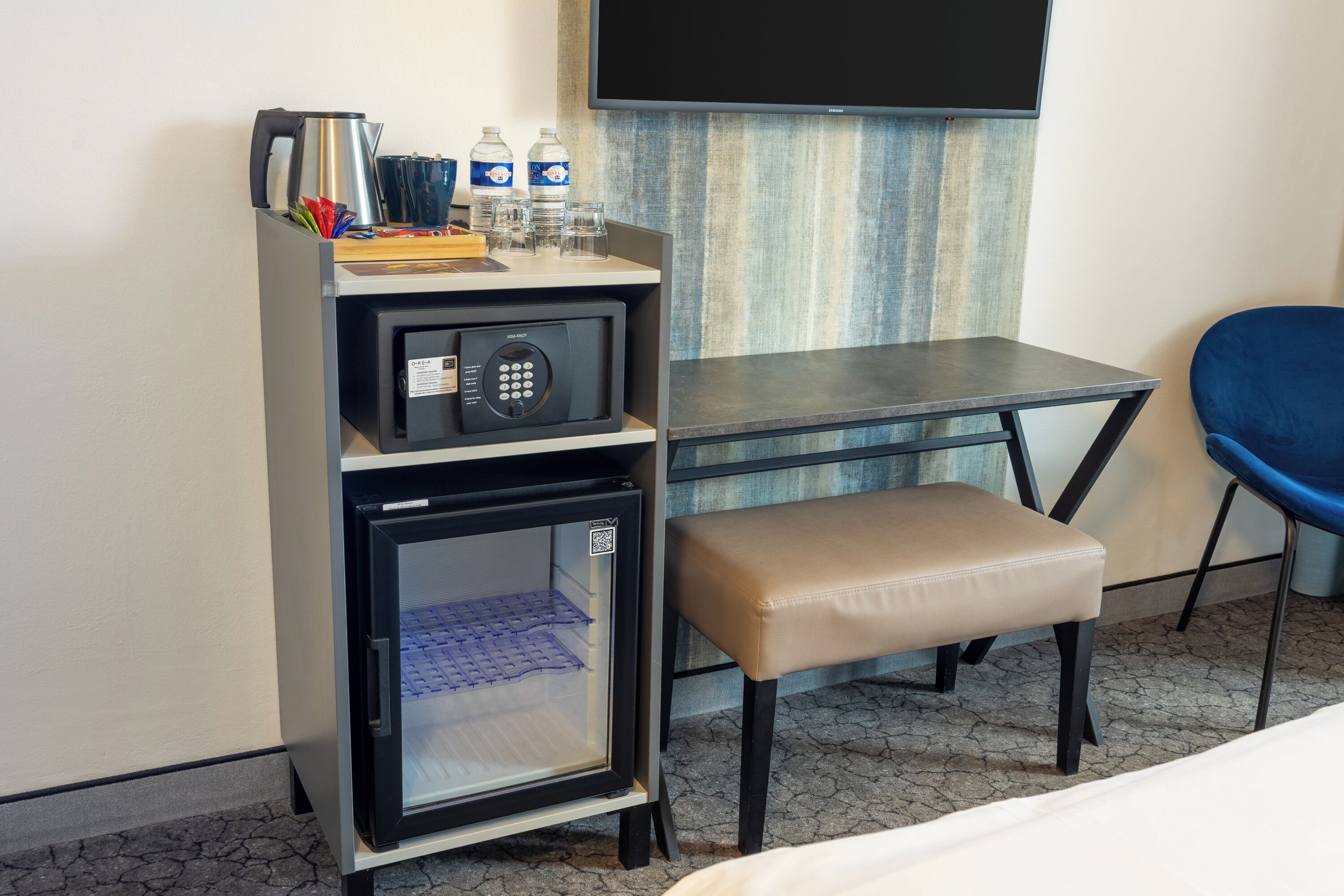 A compact hotel room with a kitchenette, a small refrigerator, a bench, a flat-screen TV, and a blue chair.