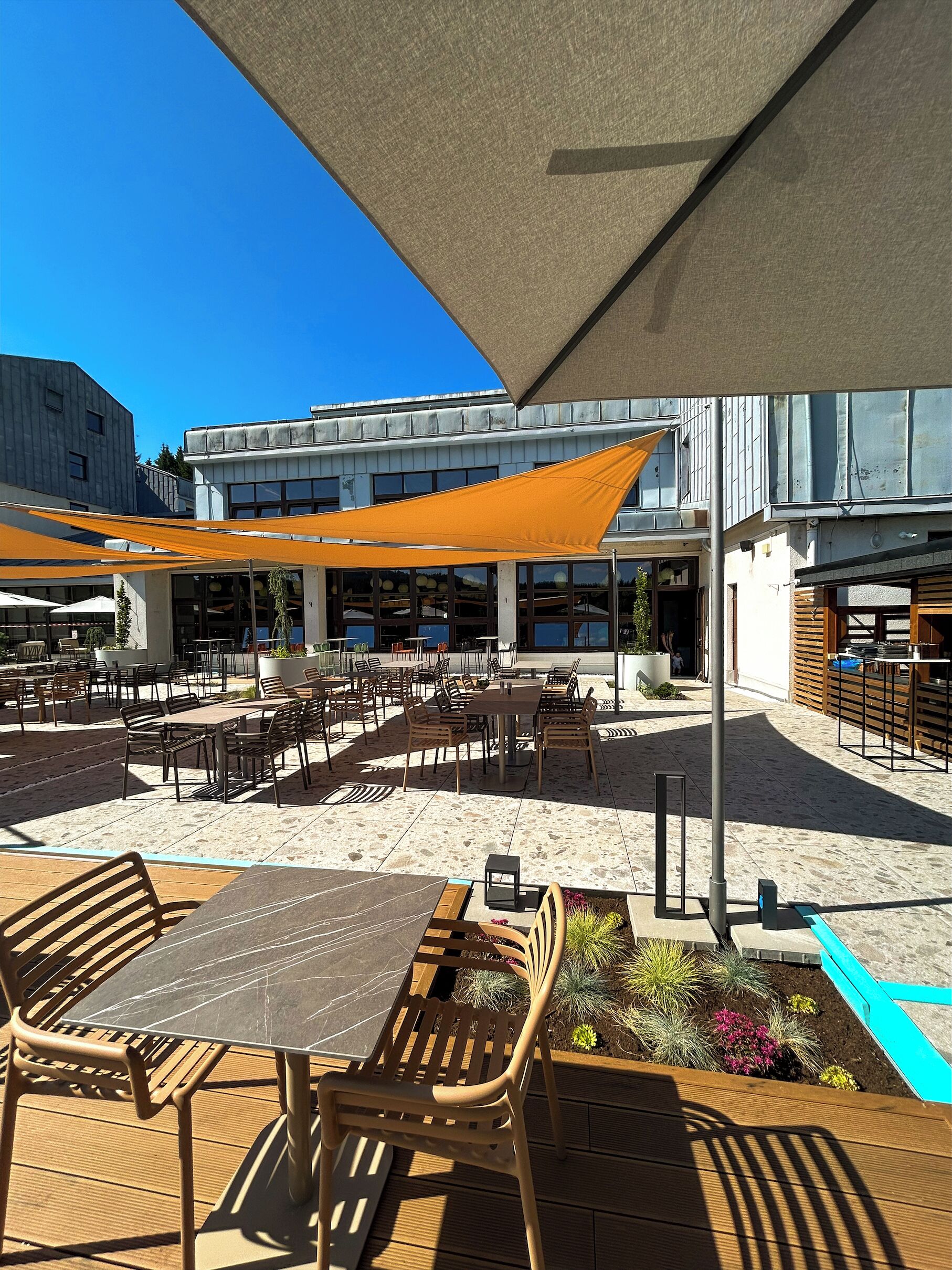 An outdoor seating area with tables and chairs, a shaded umbrella, and a view of a building with large windows.