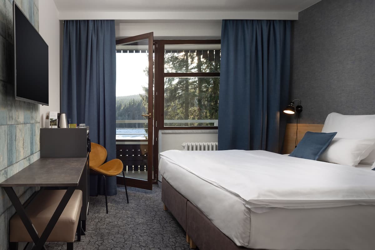 A hotel room with a bed, a desk, a chair, a TV, a window with curtains, and a view of trees.