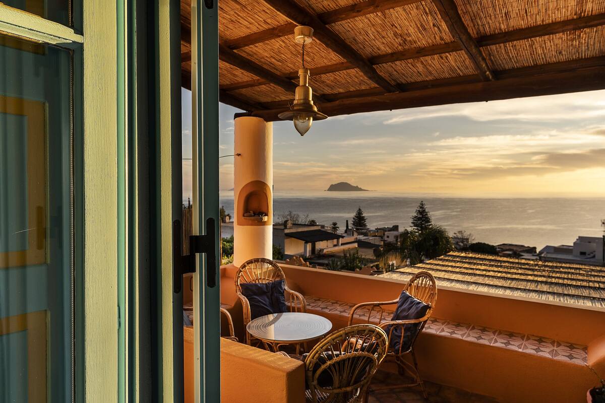 Un balcón con sillas de mimbre y una mesa, con vistas a la costa, edificios y una isla a lo lejos.
