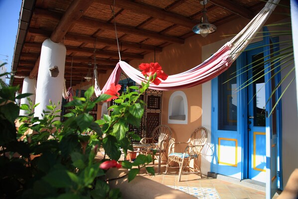 Un espacio al aire libre cubierto con sillas de mimbre, una hamaca y una puerta con detalles azules y amarillos.