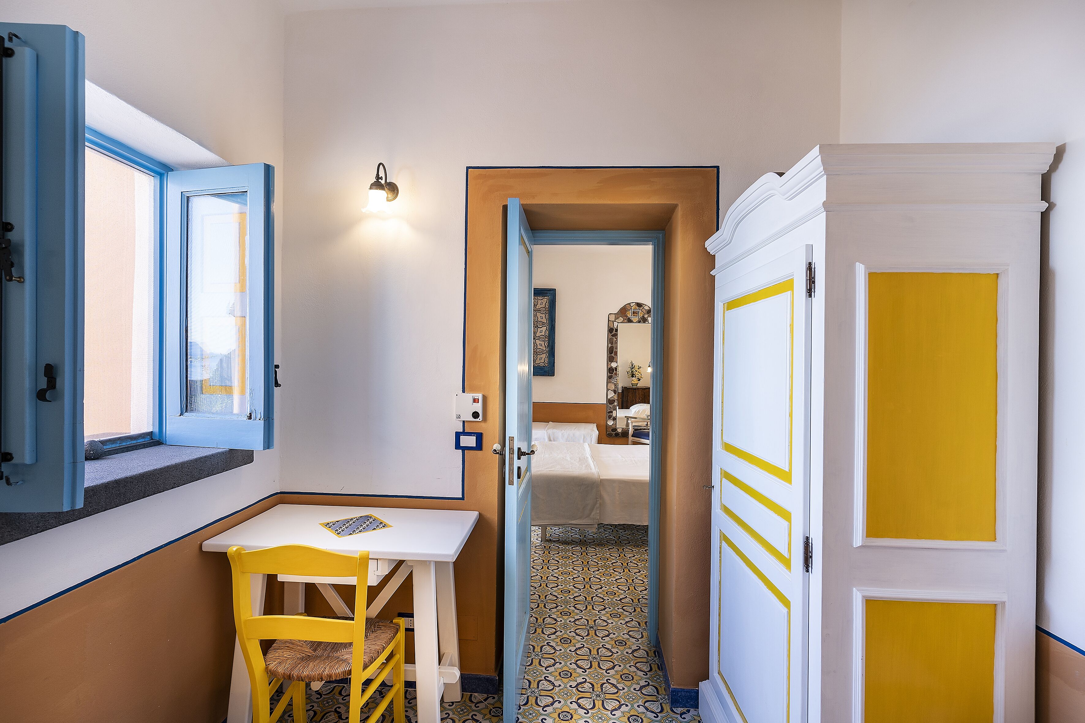 Une petite pièce comprenant une table et une chaise, une fenêtre avec des volets bleus et une porte donnant sur une autre pièce.