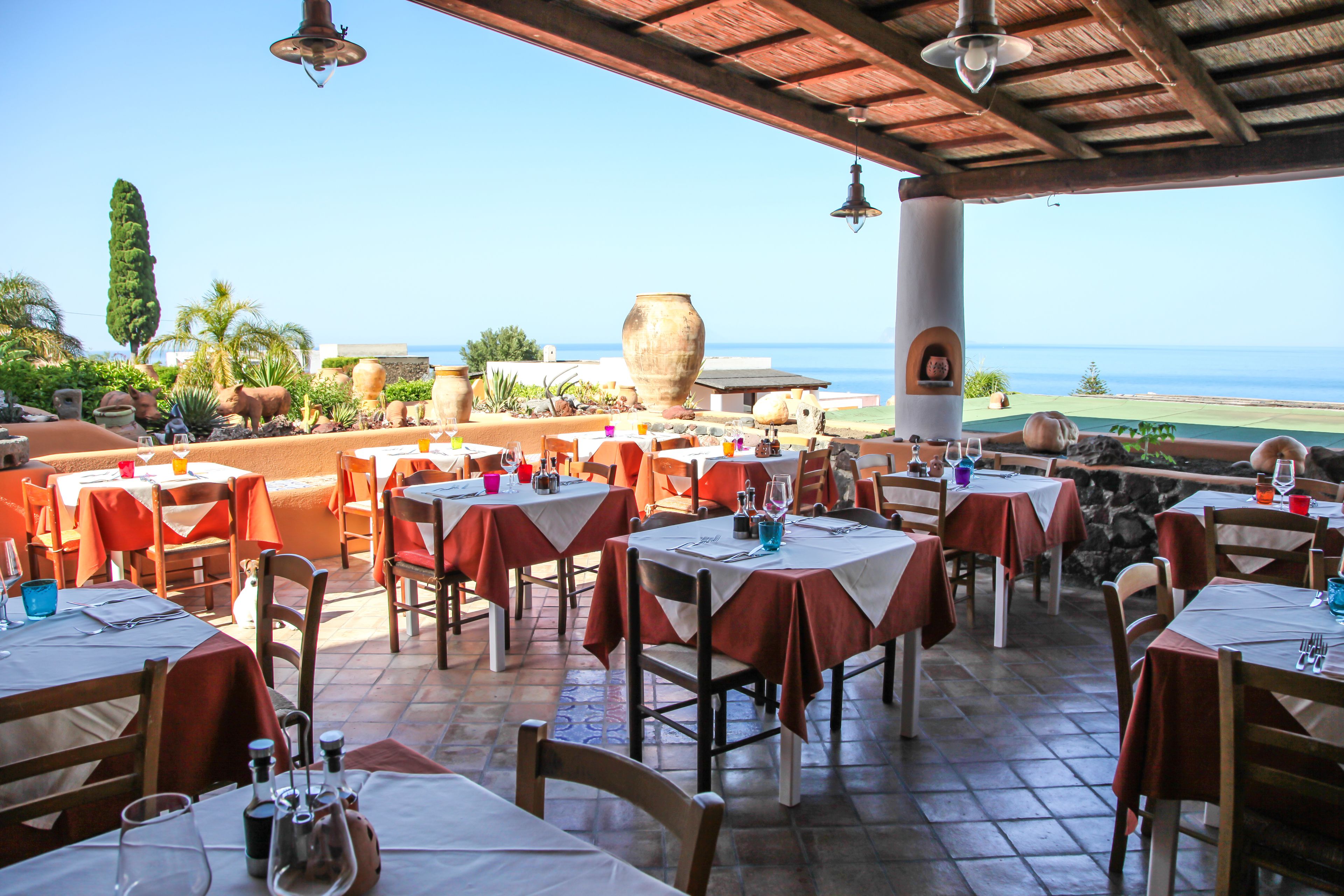 An outdoor dining area with tables and chairs, a view of the ocean, and a stone wall.