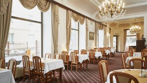 A formal dining room with multiple tables and chairs, large windows with curtains, and a chandelier.
