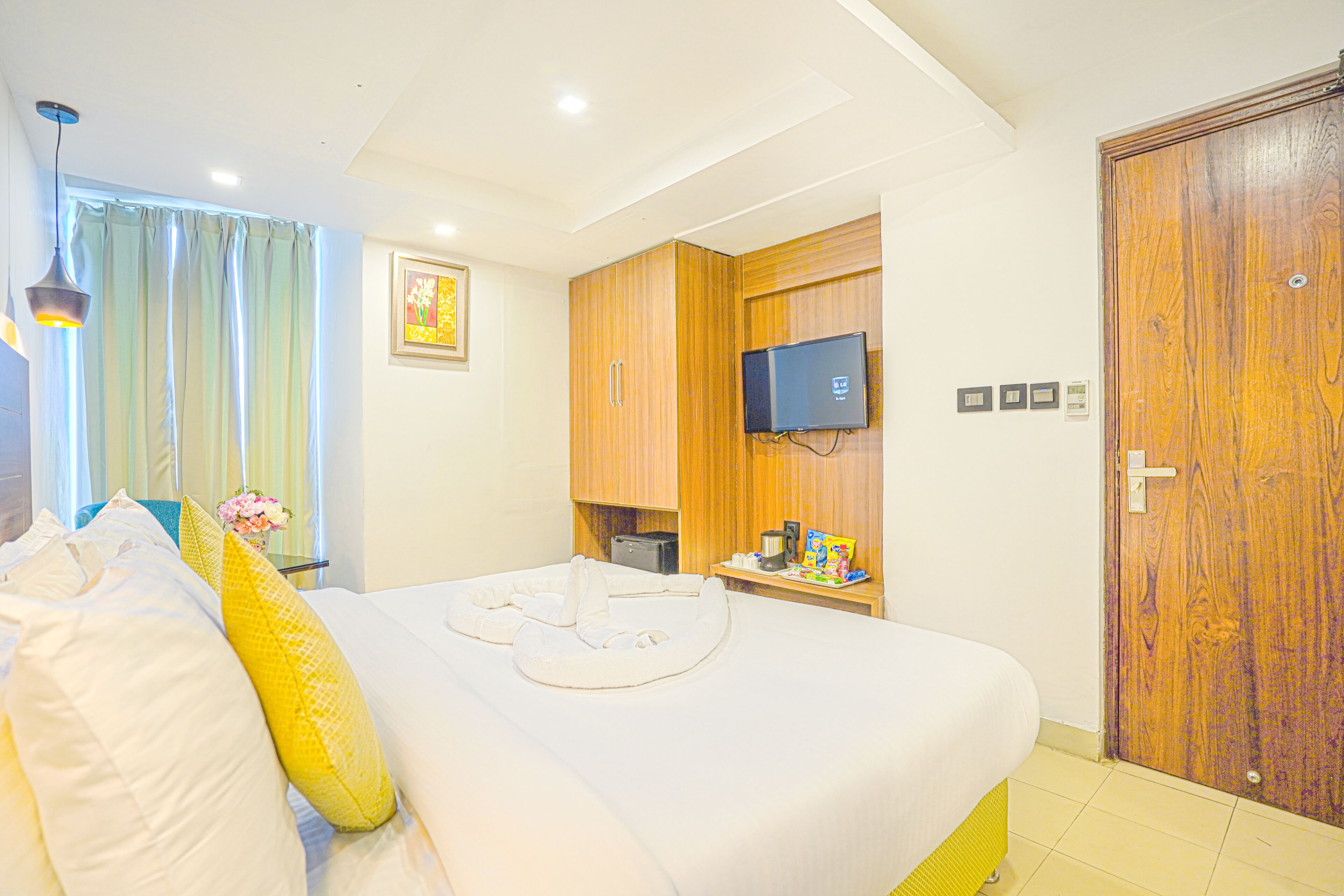 A hotel room with a large bed, a television mounted on the wall, a wooden wardrobe, and a window with curtains.