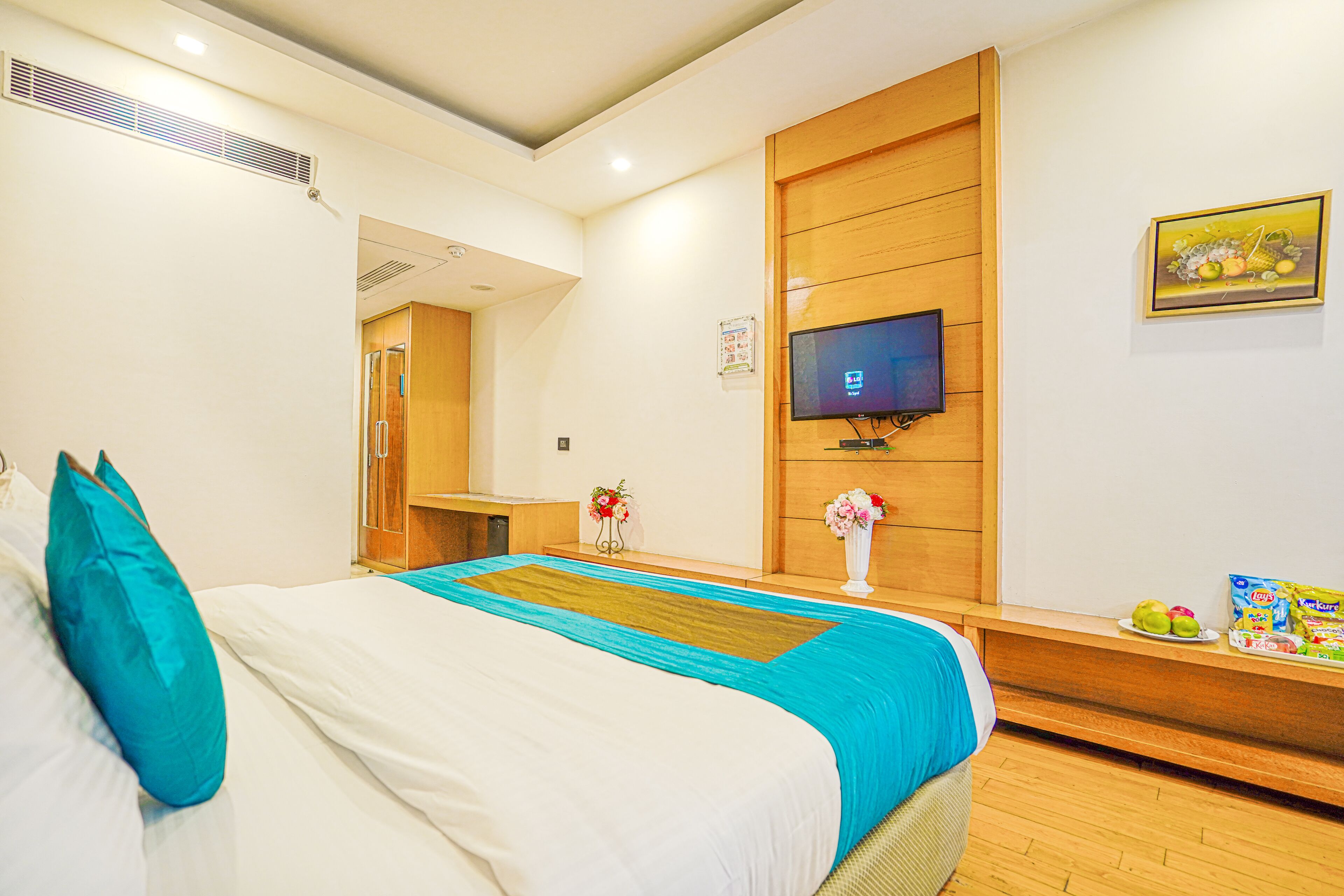A hotel room with a bed, a TV, a wooden panel wall, and a small table with fruits and snacks.