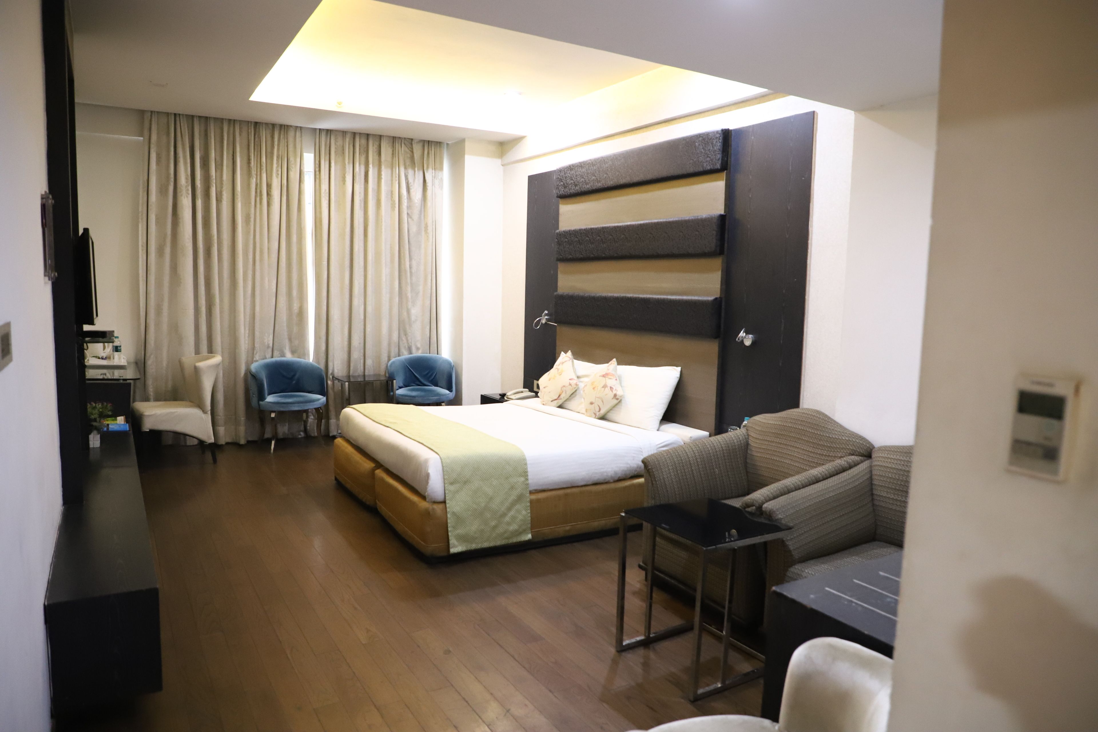 A modern hotel room with a large bed, a sofa, two armchairs, a desk, and a large window with curtains.
