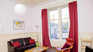 A living room with a black leather sofa, a wooden coffee table, a red leather armchair, a television, and a window with red curtains.