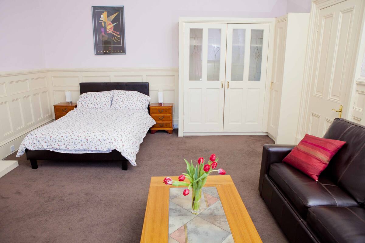 A bedroom with a bed, nightstands, a wardrobe, a coffee table with a vase of flowers, and a leather sofa.