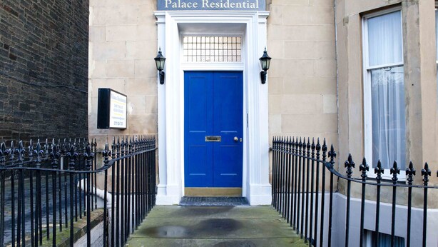 A building entrance with a blue door, a sign above reading "Palace Residential," and a black iron fence on either side.