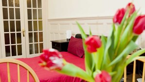 A room with a bed, a nightstand, a red pillow, a vase with red flowers, and a wooden chair.