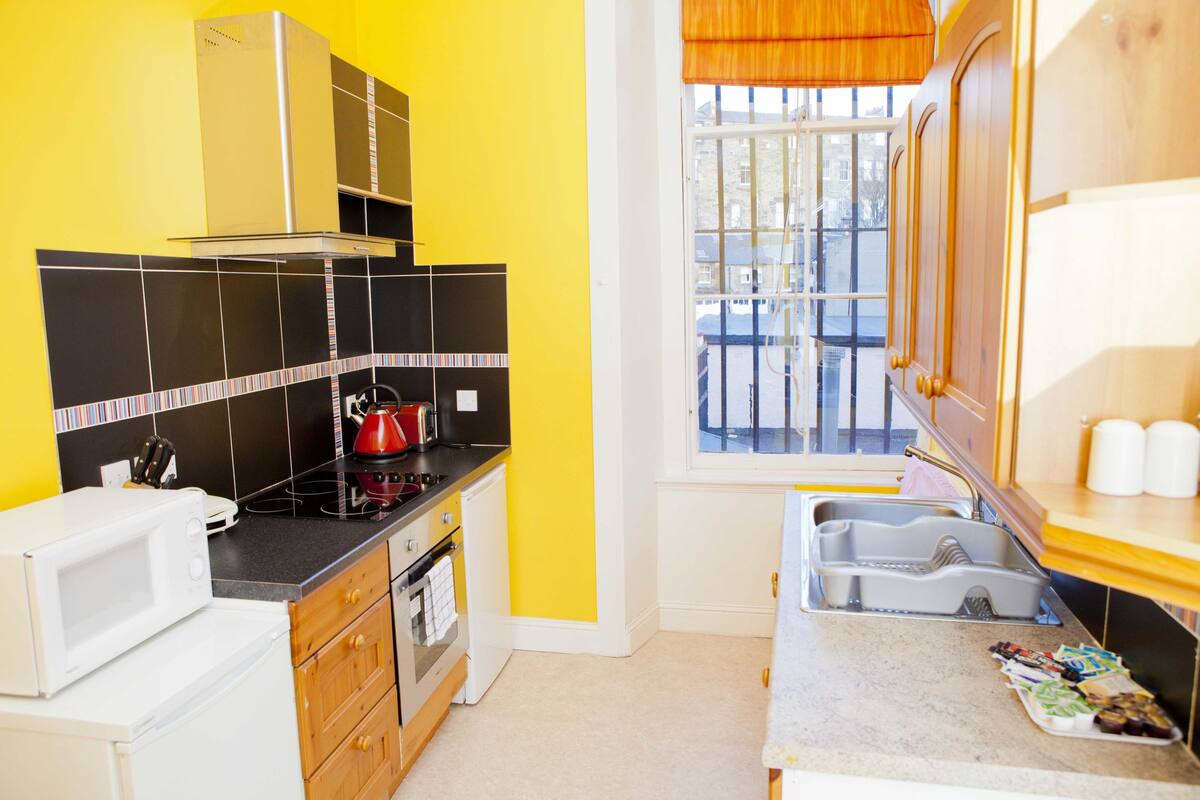 A compact kitchen with black and white tiles, a microwave, stove, and a window with a view of buildings.