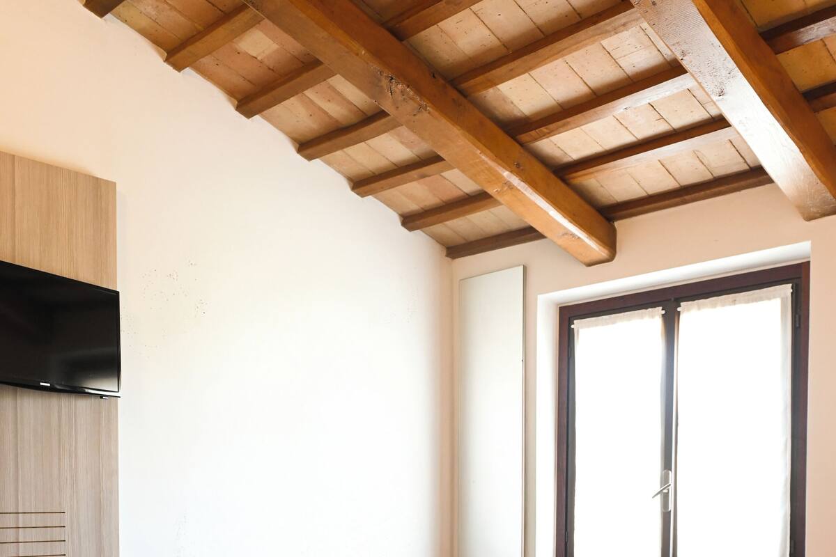 A bedroom with a bed, a bedside table with a phone, a window, and a wooden ceiling.