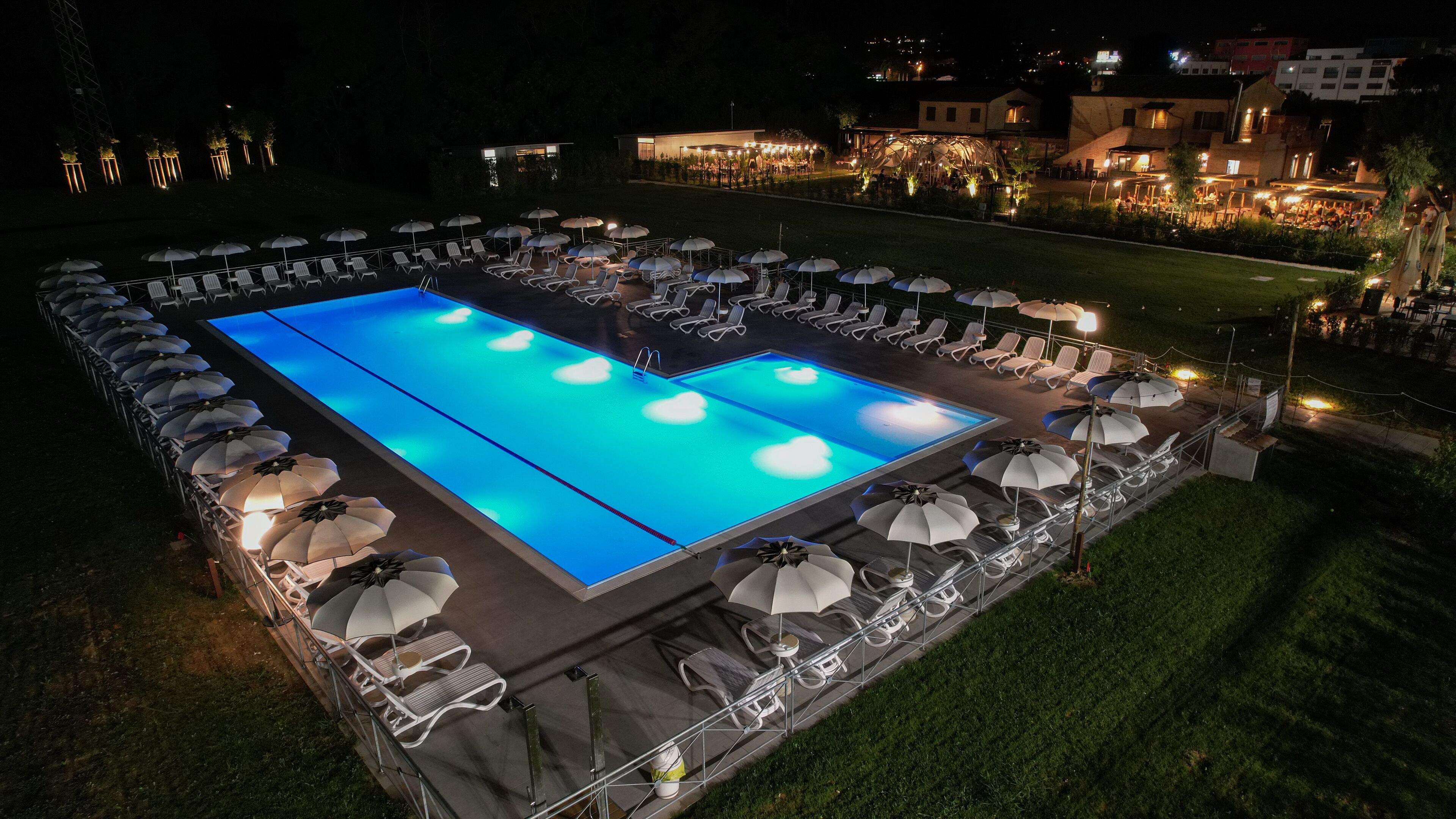 Una piscina rectangular con superficie azul iluminada, rodeada de sillas de descanso y sombrillas, con edificios a lo lejos bajo un cielo nocturno.