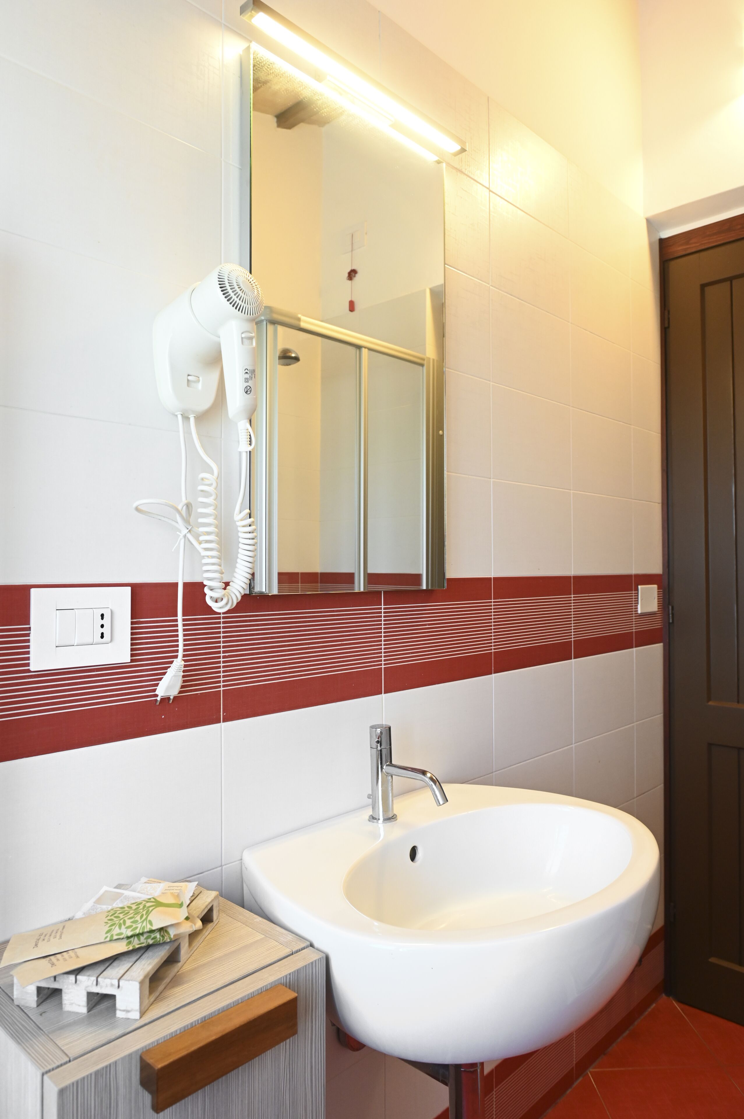 A bathroom with a white sink, a hairdryer mounted on the wall, a mirror above the sink, and a red and white tiled backsplash.