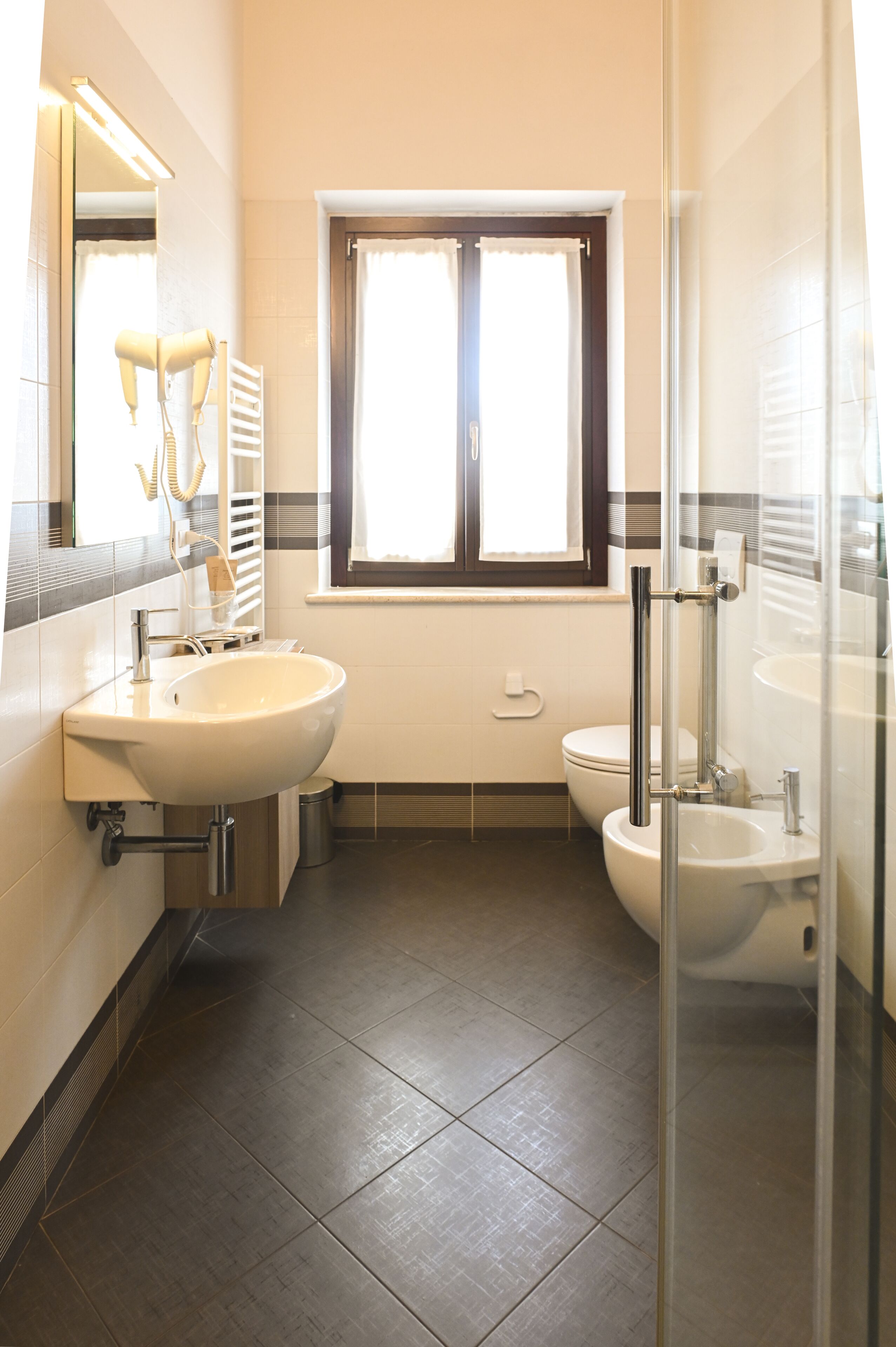 A modern bathroom with a sink, toilet, and bidet, featuring a window and a mirror.