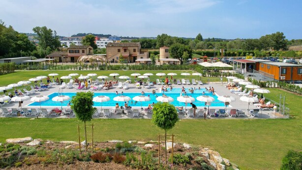 A large outdoor swimming pool surrounded by lounge chairs and umbrellas, with a view of buildings and greenery in the background.