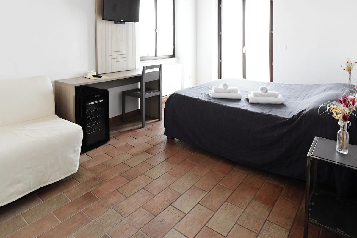 A bedroom with a bed, a desk, a chair, a TV, and a window.