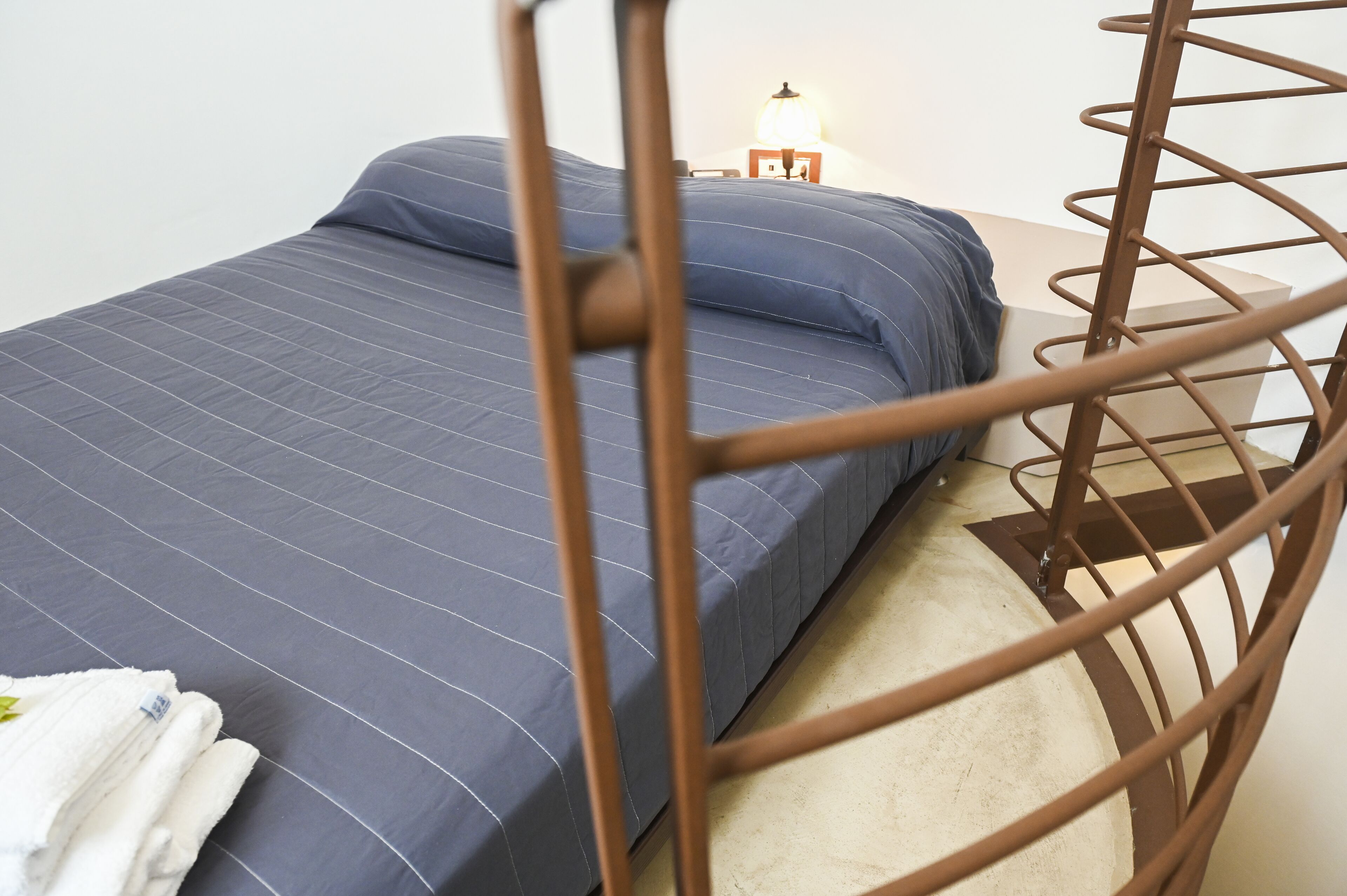 Een bed met een blauwe matras en kussens, een houten hoofdbord, en een nachtkastje met een lamp.