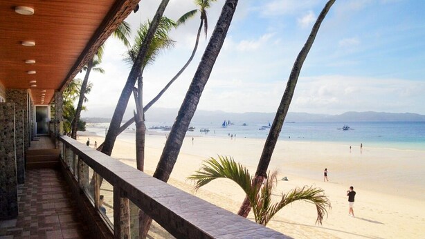 A beach view from a covered walkway with palm trees, a sandy beach, and boats in the distance. - Diamond Water Edge Resort (Boracay Island)