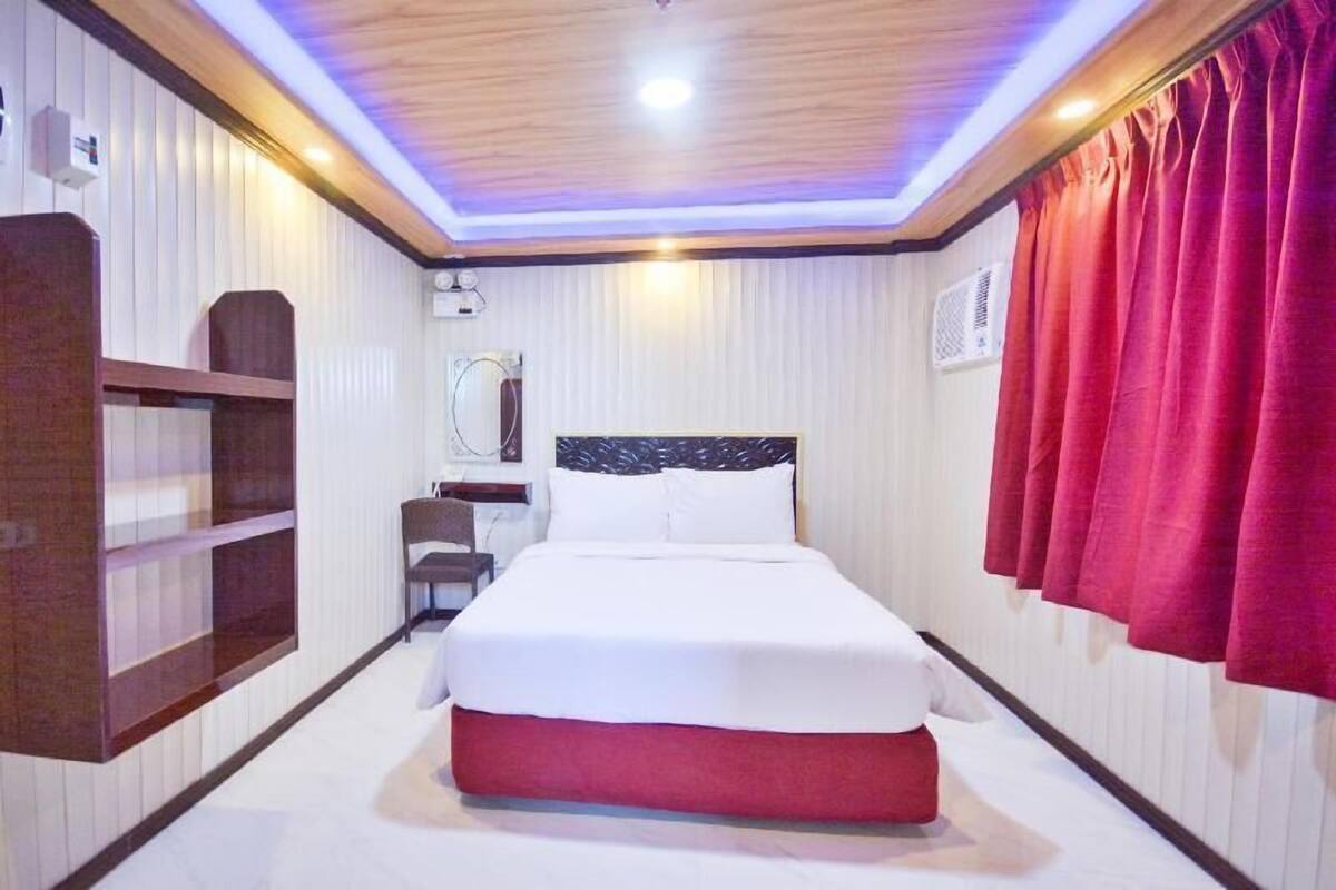A hotel room with a bed, a wooden shelf, a mirror, and red curtains.