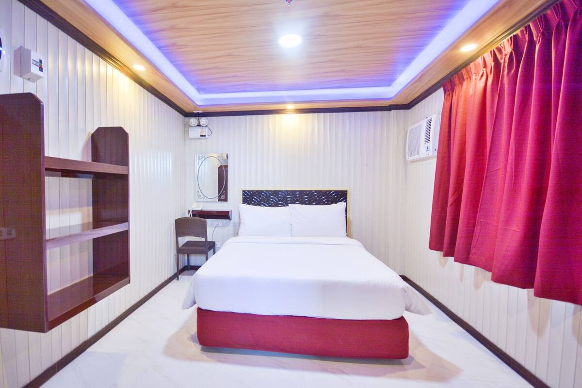 A hotel room with a bed, a wooden headboard, a red curtain, a mirror, and a shelf.