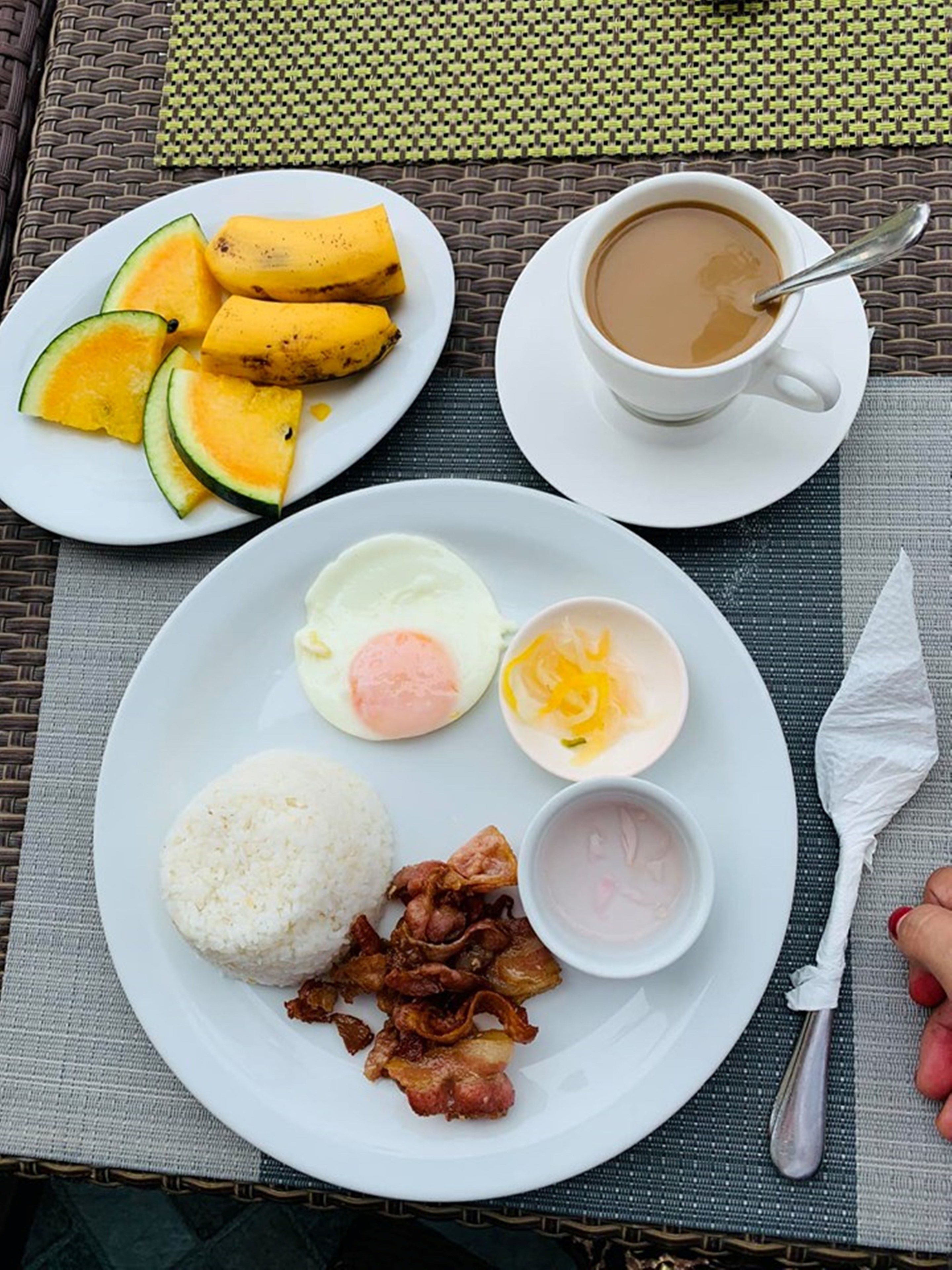 Eine Mahlzeit bestehend aus Reis, gebratenem Fleisch, einem gekochten Ei, geschnittenem Mango und einer Tasse Kaffee auf einer gewebten Tischmatte.