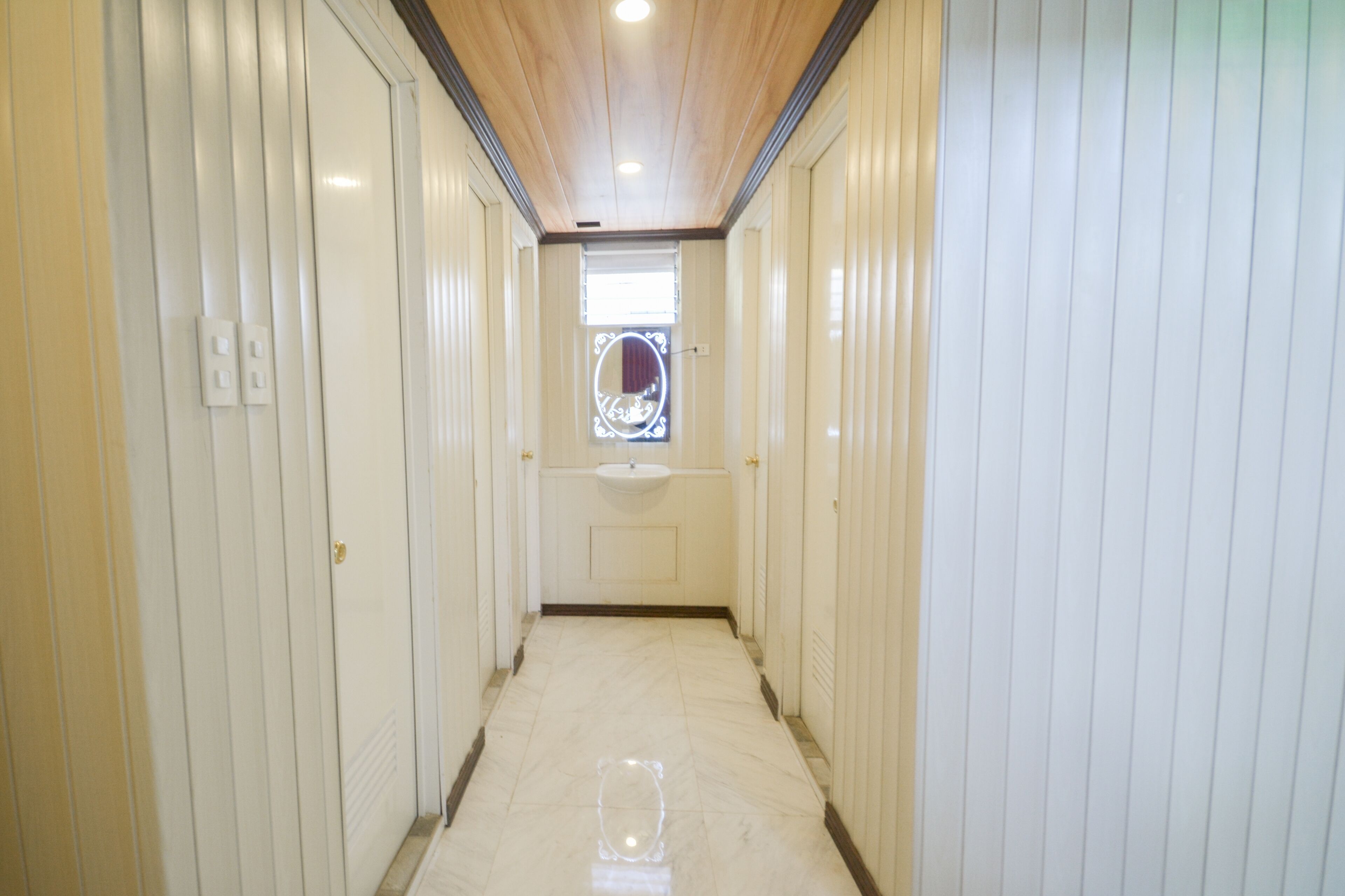 Un couloir étroit aux murs blancs et au plafond en bois, qui mène à une pièce dotée d’une fenêtre et d’un évier.