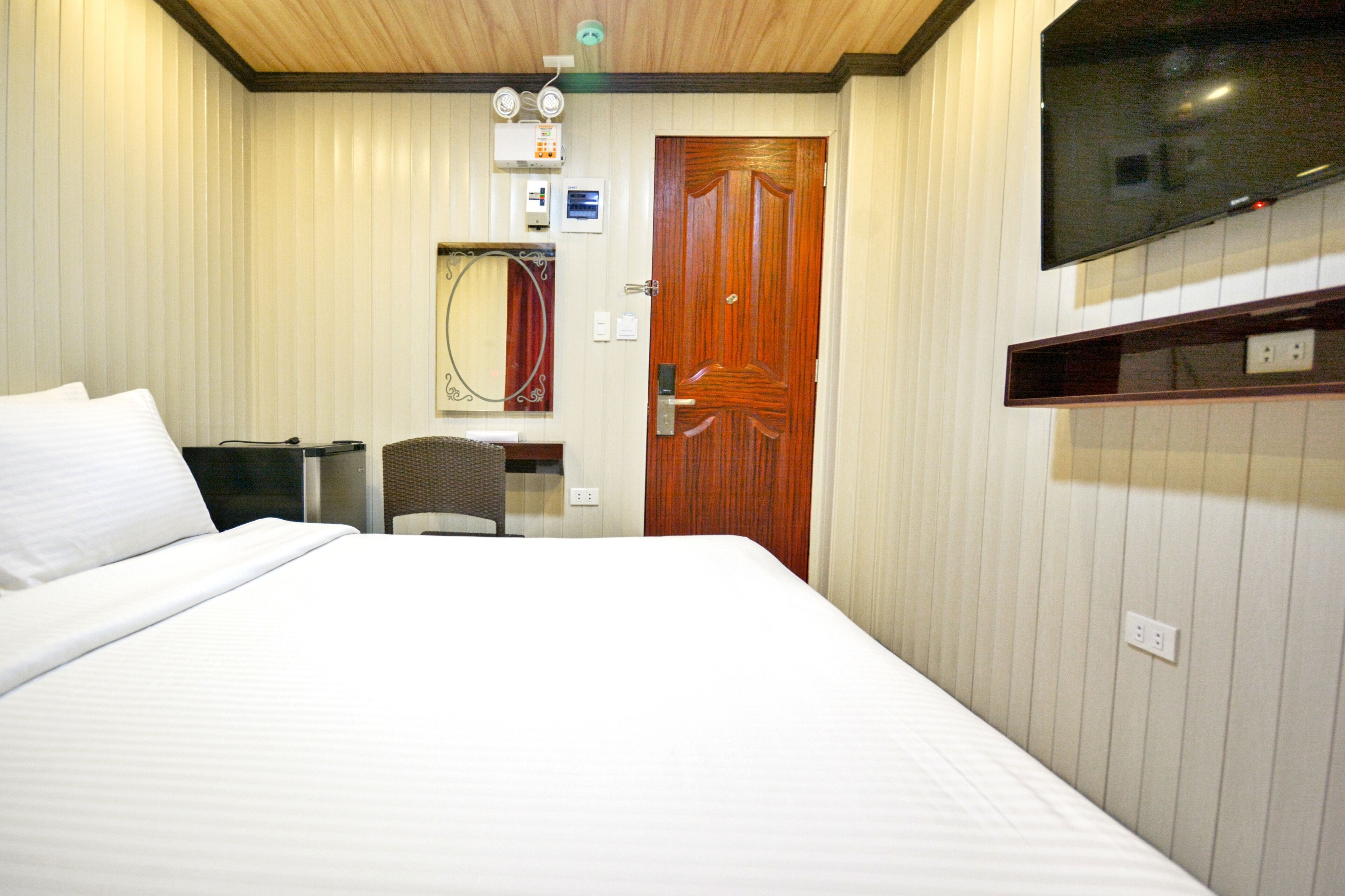 A hotel room with a bed, a wooden door, a television mounted on the wall, and a small desk with a chair.