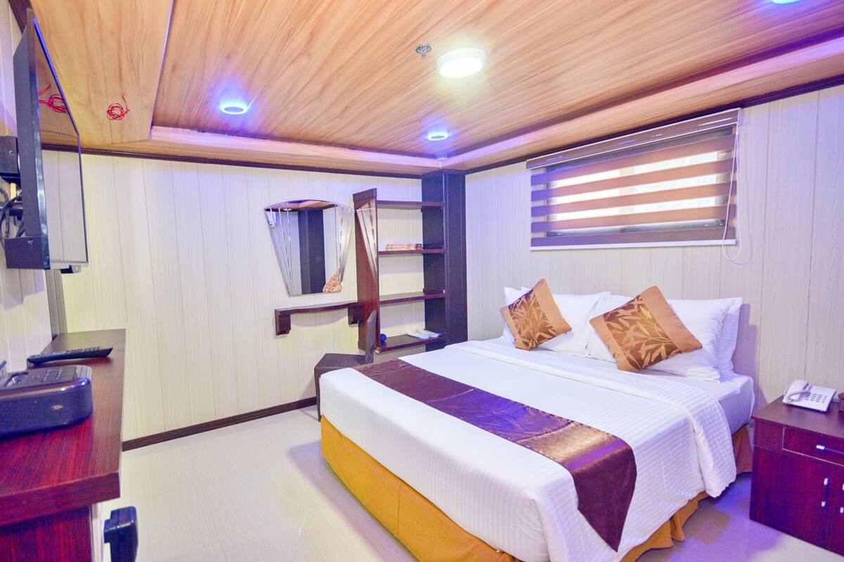 A hotel room with a wooden ceiling, a bed with white bedding and brown pillows, a bedside table with a telephone, and a window with blinds.