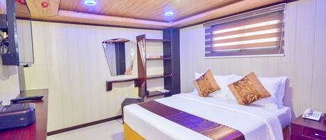 A hotel room with a wooden ceiling, a bed with white bedding and brown pillows, a bedside table with a telephone, and a window with blinds.