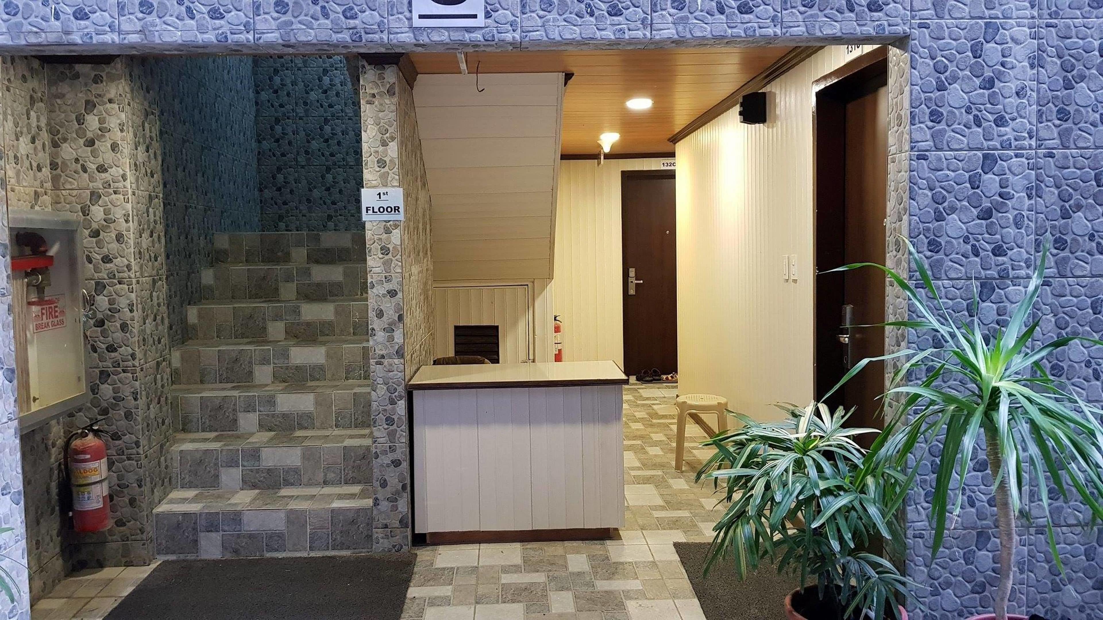 Entrada de hotel con fachada de piedra, mostrador de recepción, extintor y planta en maceta.