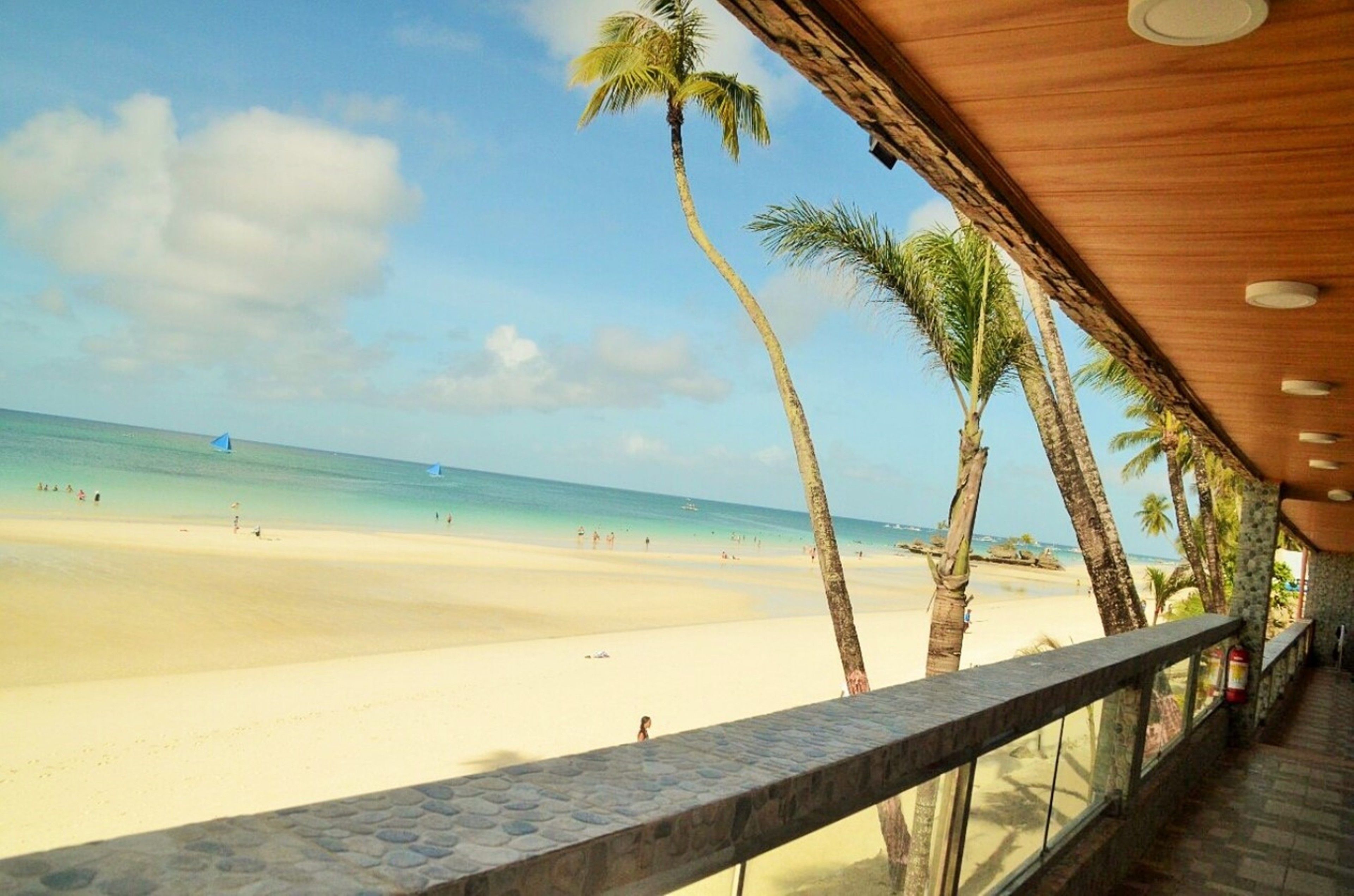 A beach view from a covered area with a wooden ceiling and stone floor, overlooking a sandy beach with palm trees and the ocean.