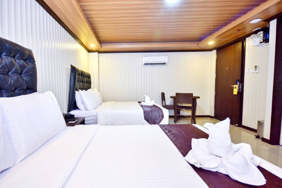 A hotel room with two beds, a wooden ceiling, an air conditioning unit, a wooden door, and a small table with a chair.