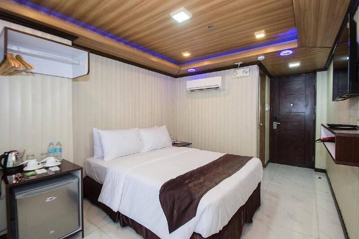 A hotel room with a bed, bedside table, air conditioning unit, and a small desk with a chair.