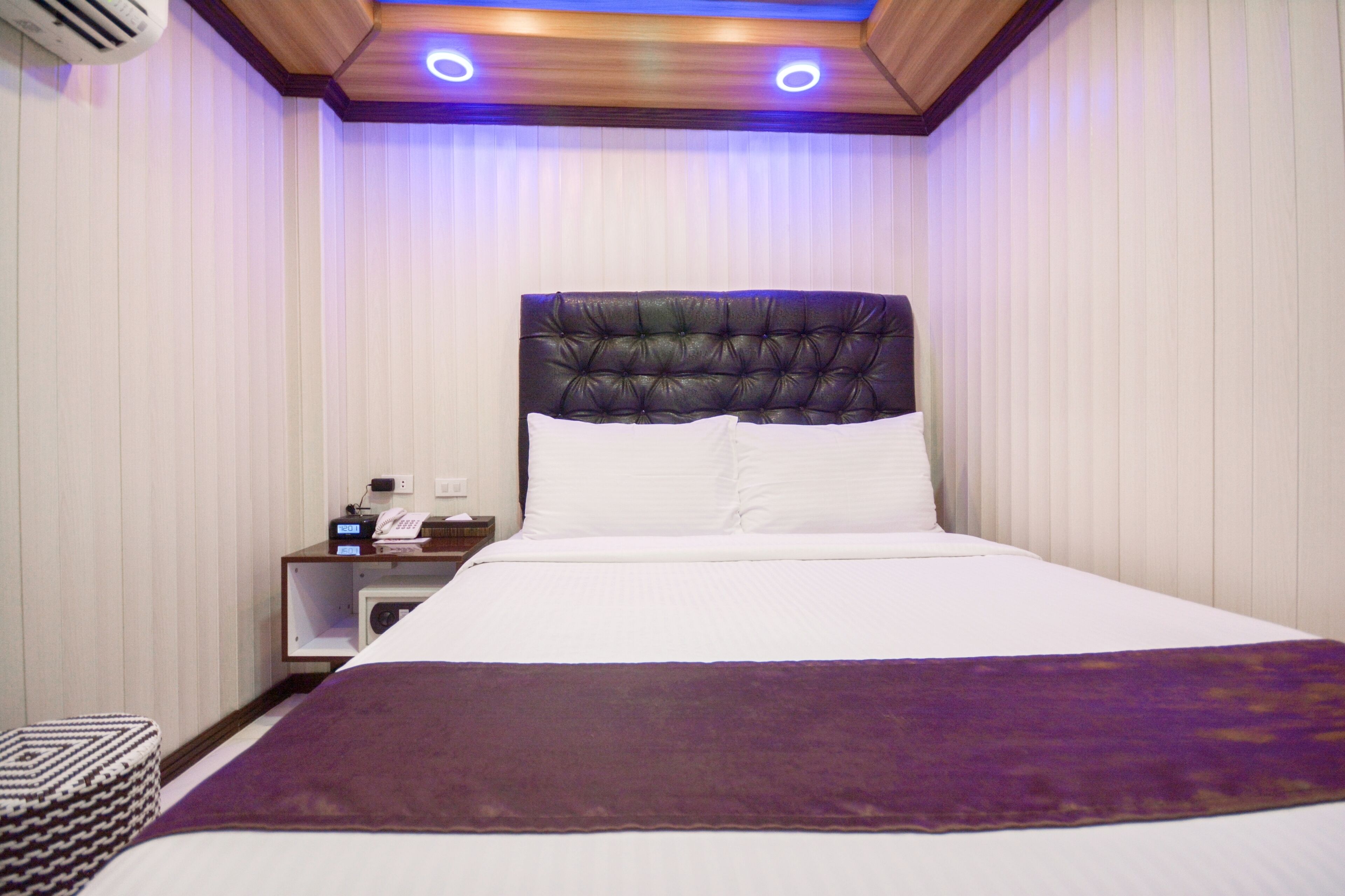 A hotel room with a large bed, a wooden headboard, a nightstand with a phone and a lamp, and a purple throw blanket.