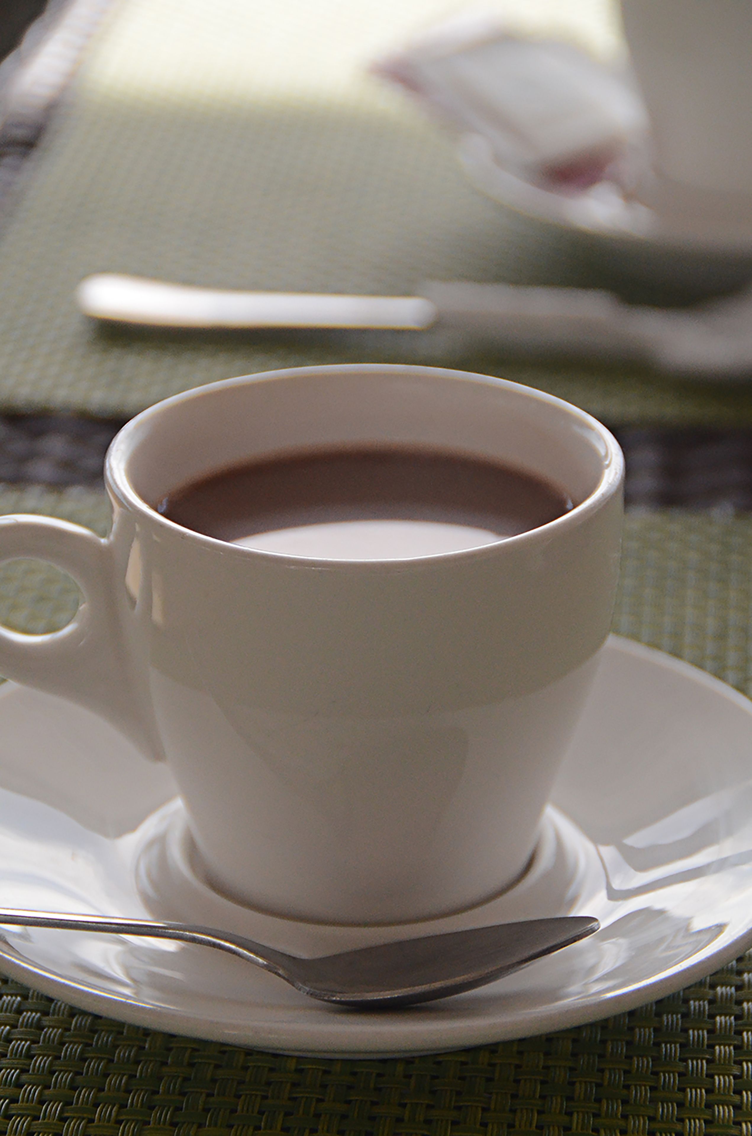 Una taza blanca de café sobre un platillo blanco con una cuchara a un lado, colocada sobre un posavasos tejido.