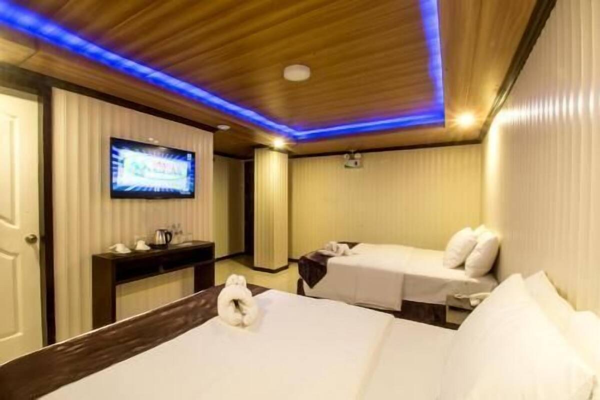 A hotel room with two beds, a television on the wall, and a wooden ceiling with blue lighting.