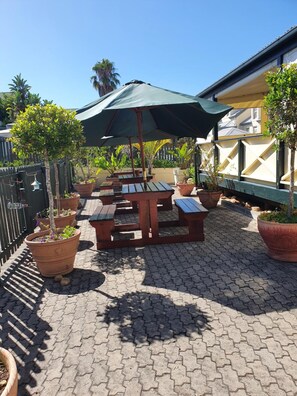 An outdoor patio with wooden tables and benches, surrounded by potted plants and a green umbrella.