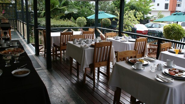 An outdoor dining area with tables set for breakfast, surrounded by greenery and a view of parked cars. - Knysna Wayside Inn (Knysna)