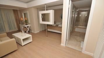 A modern hotel room with a sofa, a small coffee table, a TV mounted on a stand, a bed, and a sliding glass door leading to a shower area.