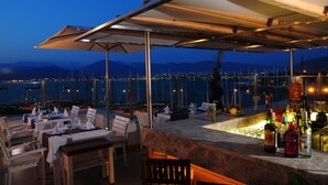 An outdoor dining area with tables and chairs, a bar with bottles, and a view of the water and mountains. - Alesta Yacht Hotel (Fethiye)