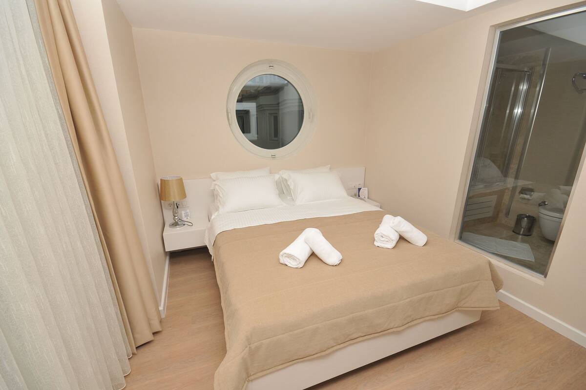 A hotel room with a bed, a round mirror, a nightstand, and a bathroom visible through a glass door.