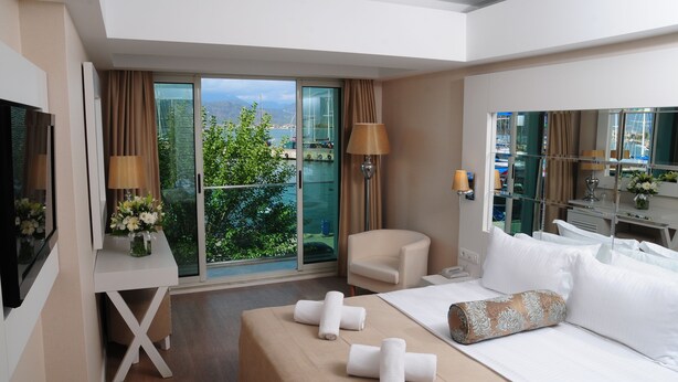 A hotel room with a bed, a desk, a chair, a TV, and a balcony with a view of the city. - Alesta Yacht Hotel (Fethiye)