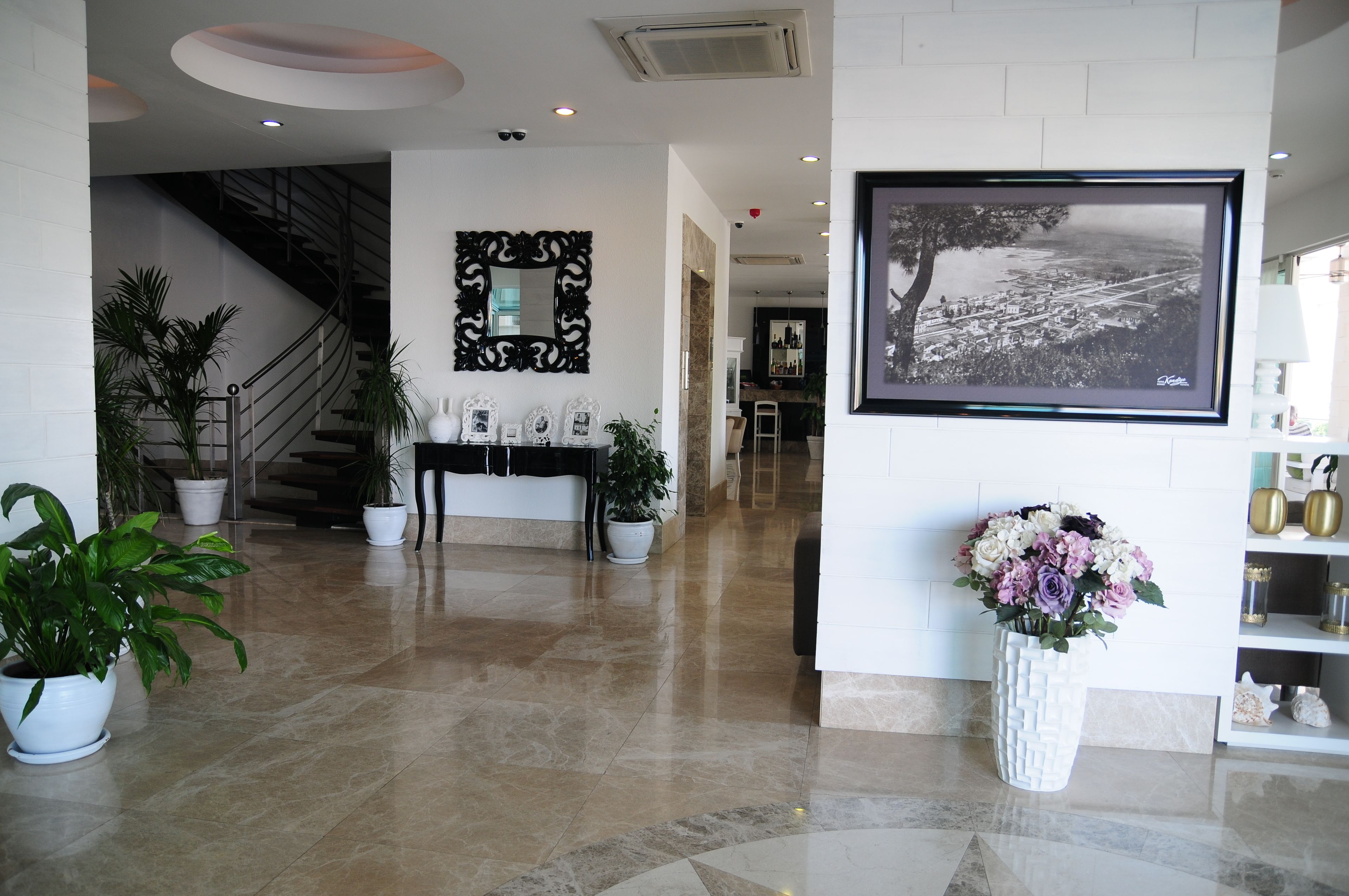 A modern lobby with a marble floor, a staircase, a reception desk, a large framed photograph, and potted plants.